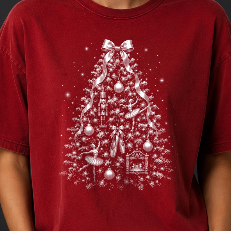 May include: A dark red t-shirt featuring a white graphic of a Christmas tree composed of ornaments, a nutcracker, ballet dancers, and ballet slippers. A large white ribbon bow sits at the top of the tree, with sparkling accents throughout the design.