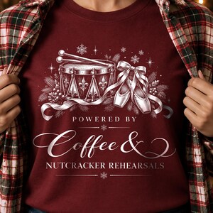 May include: A maroon t-shirt featuring a white graphic of a drum with drumsticks, ballet slippers, and pine branches. The text reads: POWERED BY Coffee & NUTCRACKER REHEARSALS. The design includes snowflake accents and decorative lines.