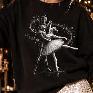 May include: A black long-sleeve sweatshirt featuring a white graphic print of a nutcracker soldier and a ballerina dancing together. The design includes swirling musical notes, staff lines, and small snowflake accents surrounding the figures.