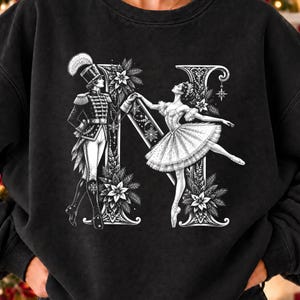 May include: A black crewneck sweatshirt featuring a large white graphic of the letter N. The letter is decorated with a nutcracker soldier and a ballerina, surrounded by intricate floral and snowflake patterns in a detailed, sketch-like style.