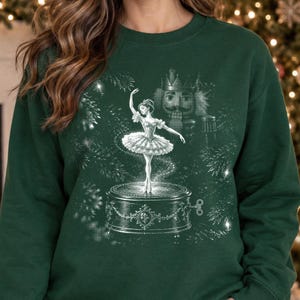 Puede incluir: Una sudadera de cuello redondo verde oscuro con un estampado gráfico blanco de una bailarina sobre una caja de música circular. Detrás de la bailarina, se observa una figura grande de cascanueces rodeada de ramas de pino y destellos.