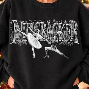 May include: A black crewneck sweatshirt featuring a white graphic print of a ballerina and a nutcracker soldier dancing. The word NUTCRACKER is written in large, stylized white letters above the figures, surrounded by a winter forest scene.