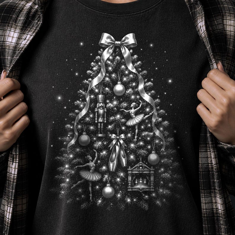 May include: A black t-shirt featuring a graphic of a Christmas tree formed by silver ornaments, including a nutcracker, two ballerinas, ballet slippers, and a small stage. A silver ribbon bow sits at the top, with sparkling lights throughout.