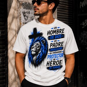 May include: A white short-sleeve t-shirt featuring a graphic print of a blue lion head next to a black cross. The text reads: UN HOMBRE DE DIOS UN PADRE ORGULLOSO UN VERDADERO PROTECTOR UN HÉROE DE VERDAD. The design uses blue and black ink.