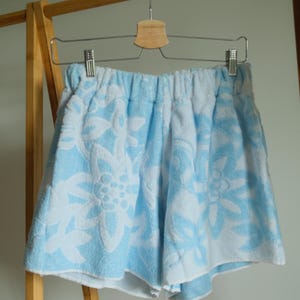 May include: Light blue and white terry cloth shorts featuring an all-over floral pattern. The shorts have an elasticized waistband and are constructed from a textured, absorbent fabric with a soft pile finish, hanging from a metal hanger.