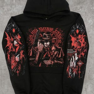 May include: A black hooded sweatshirt featuring a red and black graphic print of a character in a wide-brimmed hat holding a firearm. The design includes circular text, gothic motifs, and detailed illustrations on both sleeves with red accents.
