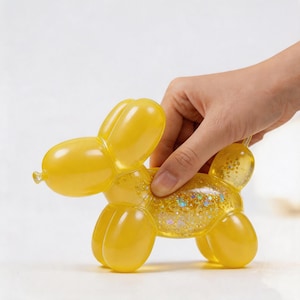 May include: A yellow balloon dog ornament with a clear body filled with glitter. The dog is held by a hand against a white backdrop. The balloon dog is a whimsical decorative item.