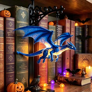 May include: A blue dragon figurine suspended from a black metal wall bracket by a chain. The dragon features detailed wings and a glowing light source within its body. It is positioned in front of a collection of vintage books and small pumpkins.