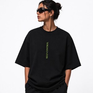 Puede incluir: Una camiseta negra de manga corta con cuello redondo, que presenta el texto "SELF-CONTROL" impreso verticalmente en una fuente de color verde lima brillante centrada en el pecho. La prenda tiene un corte holgado y cuadrado con mangas anchas.