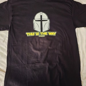 Pode incluir: Camiseta preta com um design de capacete branco e cinza com uma viseira em forma de cruz. O texto "THIS IS THE WAY" &eacute; impresso em amarelo e branco abaixo do capacete, com "(JOHN 1:5)" abaixo.