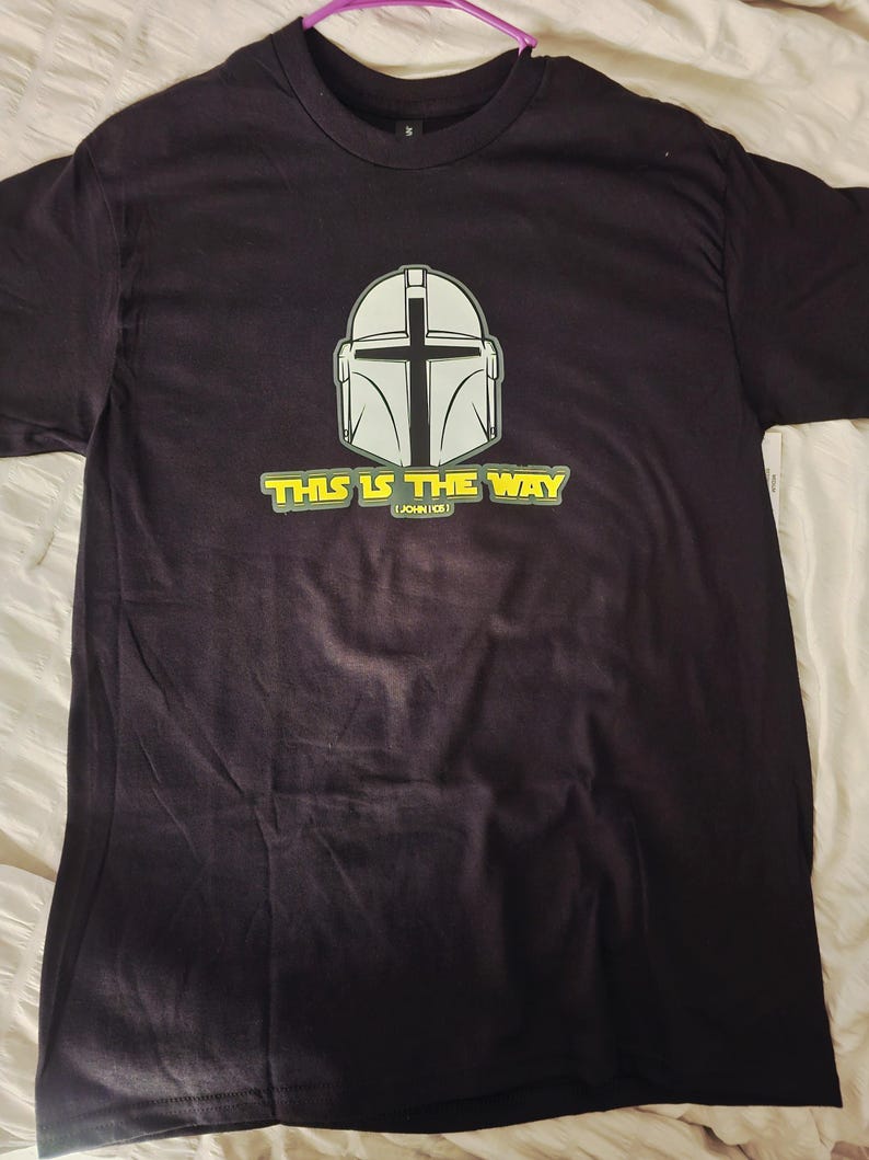 Pode incluir: Camiseta preta com um design de capacete branco e cinza com uma viseira em forma de cruz. O texto "THIS IS THE WAY" &eacute; impresso em amarelo e branco abaixo do capacete, com "(JOHN 1:5)" abaixo.