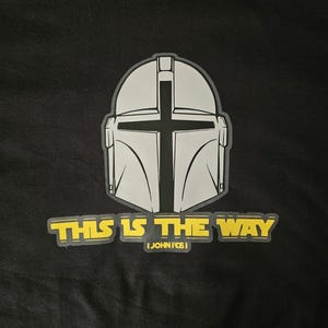 Pode incluir: Camiseta preta com um design de capacete cinza com uma cruz e o texto amarelo "THIS IS THE WAY". Abaixo est&aacute; "(JOHN 14:6)". O design est&aacute; centralizado na camiseta.