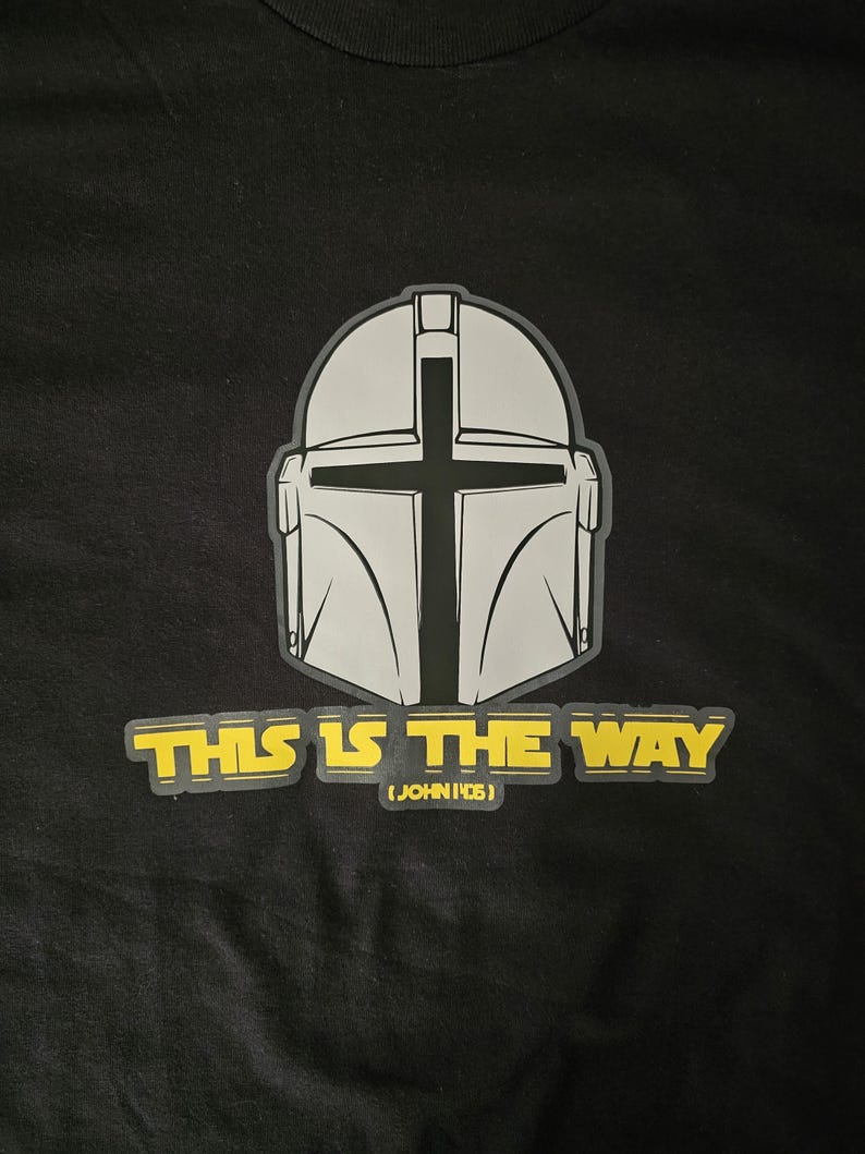 Pode incluir: Camiseta preta com um design de capacete cinza com uma cruz e o texto amarelo "THIS IS THE WAY". Abaixo est&aacute; "(JOHN 14:6)". O design est&aacute; centralizado na camiseta.