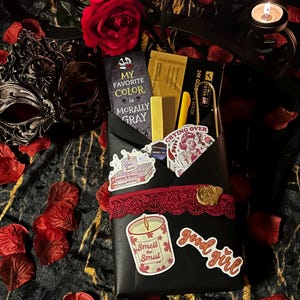 May include: A black envelope-style gift package decorated with red lace and a gold wax seal. It contains a bookmark reading MY FAVORITE COLOR IS MORALLY GRAY, a 24K gold eye mask, a yellow pen, and stickers featuring a candle and text reading good girl.