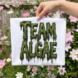 Könnte beinhalten: Ein weißes T-Shirt mit dem Text "TEAM ALGAE", gedruckt in einer dunkelgrünen, texturierten Schleim-Schriftart. Die Buchstaben haben eine moosartige, organische Oberfläche, von deren Unterseite flüssigkeitsähnliche Tropfen herabhängen.
