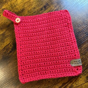 Può includere: Un sottopentola all'uncinetto rosso brillante, fatto a mano, con un passante e una chiusura a bottone. Il sottopentola ha una forma rettangolare e un'etichetta con la scritta "Made with Love by Bug."