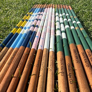 May include: A collection of wooden polo mallets with painted tips in various colors, including yellow, blue, pink, white, and green. The mallets are arranged on a bed of green grass, with the text "GRAIN AIM CO" visible on the wooden shafts.