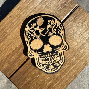 May include: A black and brown wooden sugar skull ornament with intricate floral designs. The skull has a hole at the top for hanging.