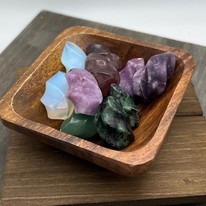 May include: A wooden bowl filled with various polished stones in shades of purple, green, and white. The stones are arranged in a random pattern.