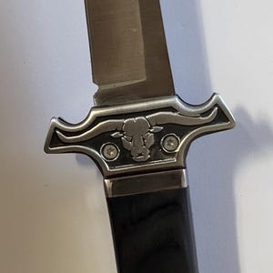 May include: A fixed-blade knife featuring a silver-toned metal guard embossed with a bull head design and two visible screws. The handle is made of dark, smooth wood with two metal rivets, and the pommel displays an intricate silver floral pattern.