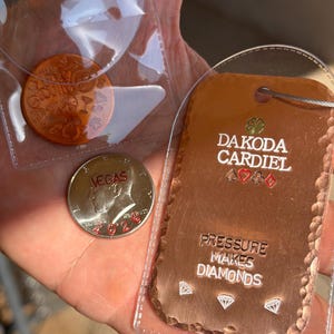 May include: A copper-colored rectangular tag with the text DAKODA CARDIEL, a clover, card suits, and the phrase PRESSURE MAKES DIAMONDS with diamond icons. Beside it is a silver-colored coin marked VEGAS 2023 and a round copper token in a plastic sleeve.