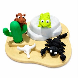 Puede incluir: Una pieza decorativa con temática desértica que presenta un cactus verde, un animal marrón, una calavera blanca, una rana verde y un escorpión negro. Las figuras están dispuestas sobre una base beige texturizada.