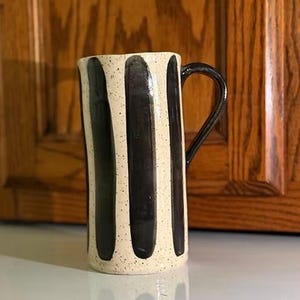 May include: A tall ceramic mug featuring a cream-colored base with a speckled texture and vertical black stripes. The mug has a cylindrical body and a single curved black handle attached to the side, providing a contrast to the light-colored surface.