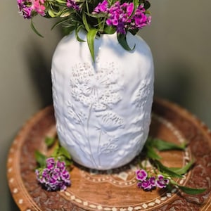May include: A white ceramic vase with embossed floral patterns displays a bouquet of pink and purple flowers. The vase is placed on a carved wooden table, creating a natural and elegant display. The flowers add a splash of colour.