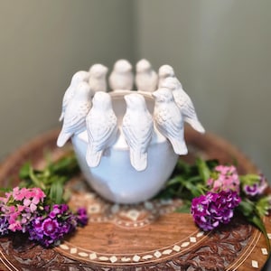 May include: A white ceramic bowl with bird figurines around the rim. The bowl is on a carved wooden tray with floral designs, surrounded by pink and purple flowers. The tray is made of wood.