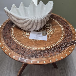 May include: A white ceramic clam shell bowl with a ribbed exterior texture. The bowl sits on a round wooden table featuring intricate floral carvings and inlaid diamond-shaped accents. A small white card displays the text the hedgehog next door.