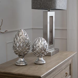 May include: Two silver-coloured decorative pinecone-shaped ornaments with layered, pointed scales, each mounted on a circular pedestal base. The ornaments are positioned in front of a tall, rectangular silver lamp with a grey fabric lampshade.