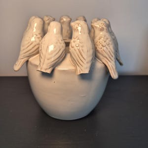 May include: A cream-coloured ceramic flower frog or vase featuring a circular arrangement of eight small, sculpted birds perched along the rim. The birds have textured wing details and a glossy finish, while the base is a smooth, rounded vessel.