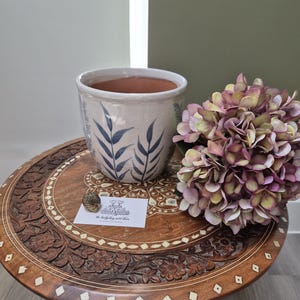 May include: A ceramic planter with a white glaze and blue leaf patterns, a cluster of purple and green hydrangea flowers, and a small business card with the text "the hedgehog next door" and a tiny metallic hedgehog figurine on top.