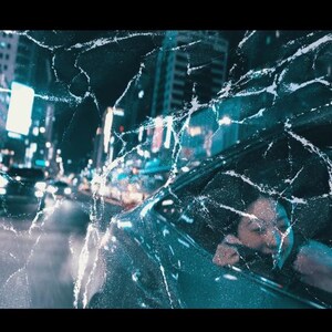 May include: A nighttime cityscape with bright lights and moving vehicles, viewed through a cracked, textured surface. A person is visible in a car, looking out the window. The image has a cinematic, urban theme.
