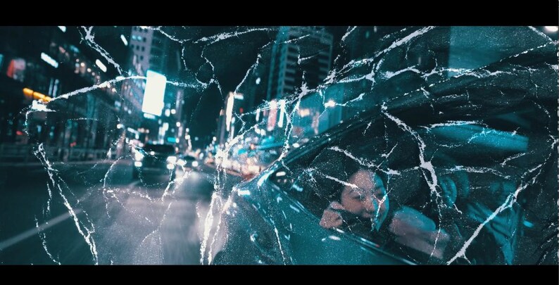 May include: A nighttime cityscape with bright lights and moving vehicles, viewed through a cracked, textured surface. A person is visible in a car, looking out the window. The image has a cinematic, urban theme.