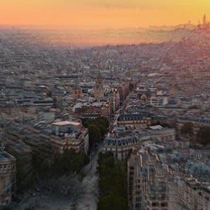 May include: Aerial view of a cityscape at sunset. Buildings with light-colored facades line the streets, with a prominent church visible in the distance. The sky transitions from orange to a soft yellow.