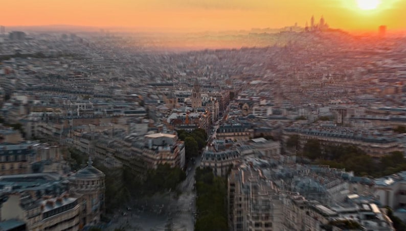 May include: Aerial view of a cityscape at sunset. Buildings with light-colored facades line the streets, with a prominent church visible in the distance. The sky transitions from orange to a soft yellow.