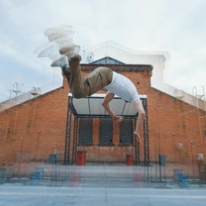 May include: A person performing a backflip in front of a brick building. The person is wearing a white t-shirt and khaki pants. The sky is overcast.