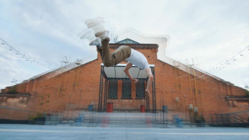 May include: A person performing a backflip in front of a brick building. The person is wearing a white t-shirt and khaki pants. The sky is overcast.