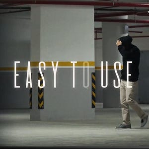 以下が含まれることがあります： 駐車場を背景に「EASY TO USE」の文字が書かれた画像。黒いパーカーとカーキ色のズボンを履いた人物がカメラから離れて歩いています。コンクリートの柱と黄黒の安全標識があり、薄暗いシーンです。