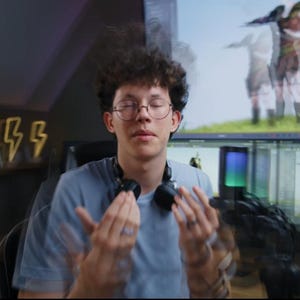 May include: A person wearing glasses and a light blue t-shirt with headphones around their neck. The person is in front of a computer monitor displaying a 3D rendering. Decorative lightning bolt lights are on the wall.
