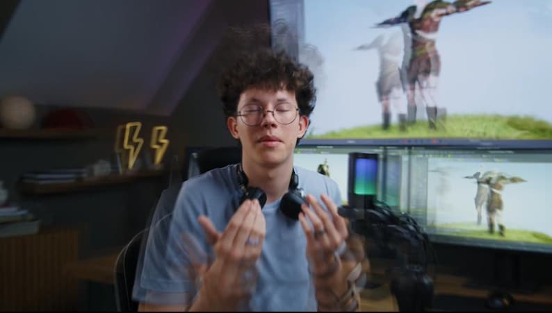 May include: A person wearing glasses and a light blue t-shirt with headphones around their neck. The person is in front of a computer monitor displaying a 3D rendering. Decorative lightning bolt lights are on the wall.