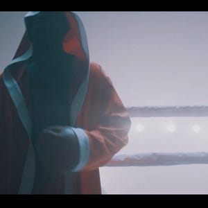May include: A red boxing robe with white trim and a hood is worn by a person in a boxing ring. The person is wearing black boxing gloves. The boxing ring has white ropes and is lit by overhead lights.