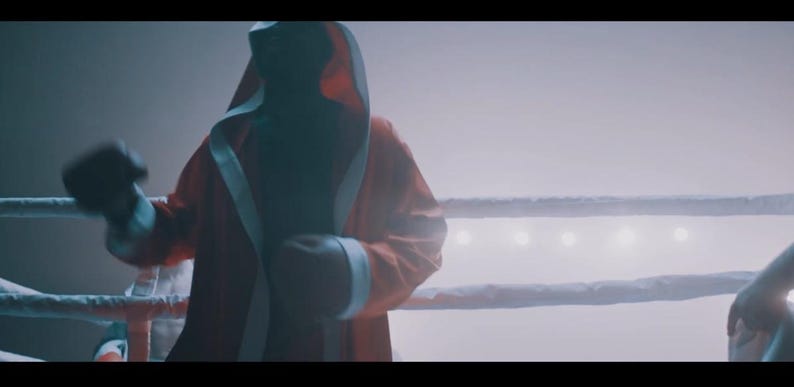 May include: A red boxing robe with white trim and a hood is worn by a person in a boxing ring. The person is wearing black boxing gloves. The boxing ring has white ropes and is lit by overhead lights.
