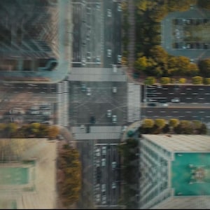 May include: Aerial view of a city intersection with crosswalks, roads, and buildings. The image has a blurred effect, suggesting motion. Cars and trees are visible, with a focus on the urban landscape.