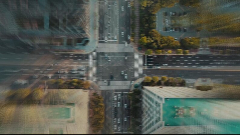 May include: Aerial view of a city intersection with crosswalks, roads, and buildings. The image has a blurred effect, suggesting motion. Cars and trees are visible, with a focus on the urban landscape.