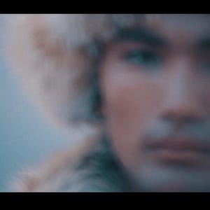 May include: A person wearing a fur hat, with a blurred background. The person's face is in focus, with a neutral expression. The hat appears to be made of light-colored fur.