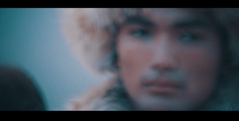 May include: A person wearing a fur hat, with a blurred background. The person's face is in focus, with a neutral expression. The hat appears to be made of light-colored fur.