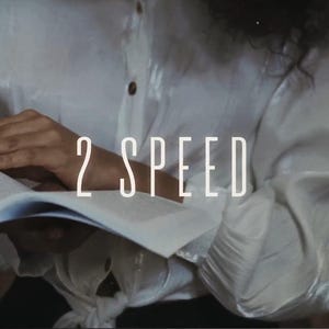 以下が含まれることがあります： 「2 SPEED」の文字が重ねられた、開いた本を持つ人物。白いボタンダウンシャツを着ています。背景はぼやけた本棚です。ウェブサイトのナビゲーションが上部に表示されています。