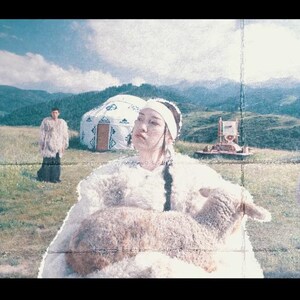 May include: A pastoral scene featuring people in traditional fur garments, standing near white yurts on a grassy field. One person holds a lamb. Mountains and a blue sky with clouds are in the background.